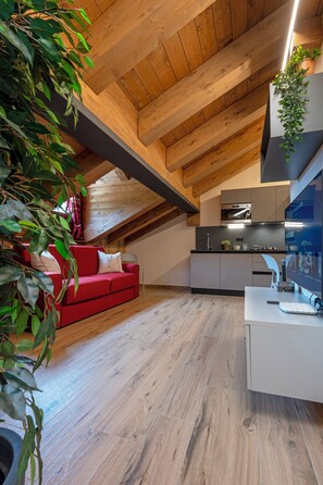 Ein modernes Loft mit freiliegenden Holzbalken, einem roten Sofa, einer Küche mit grauen Schränken und einem großen Fenster.