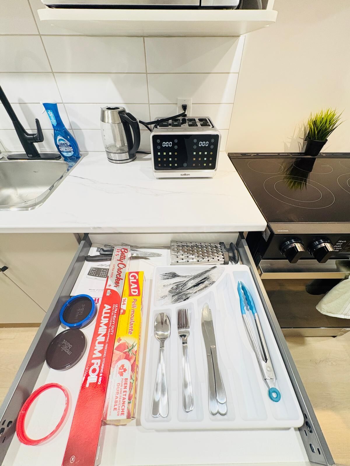 Una cocina con un reloj digital, un hervidor, un tostador y un cajón con utensilios de cocina y papel de aluminio.