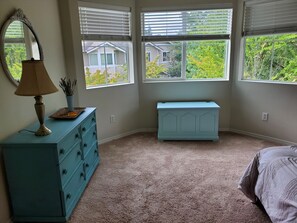 A bedroom with a dresser, a lamp, a bench, and a view of the outside.