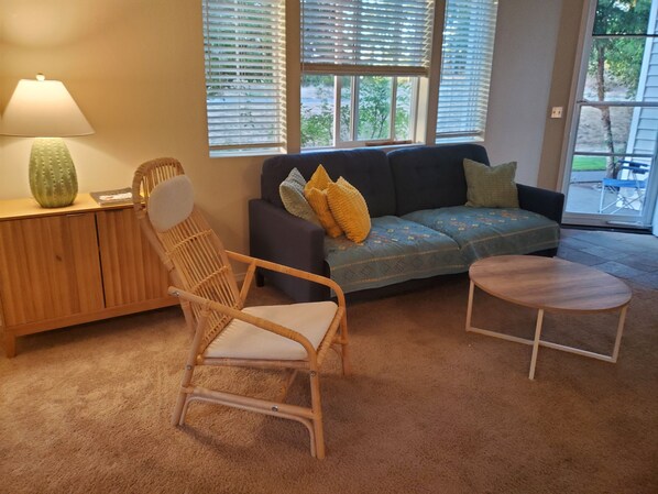 A living room with a wicker chair, a sofa with patterned cushions, a small round table, and a lamp on a wooden cabinet.