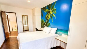 Quarto de hotel com cama, escrivaninha, cadeira, guarda-roupa e parede com um mural tropical.