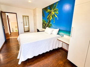 Une chambre d’hôtel comprenant un lit, un bureau, une chaise, une armoire et un mur orné d’une fresque aux motifs tropicaux.