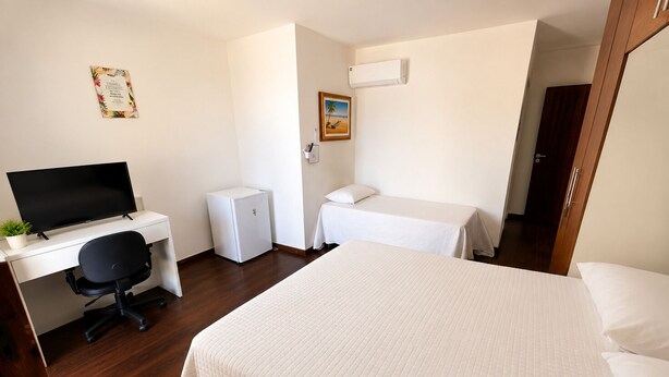 Quarto de hotel com cama, escrivaninha com computador, mini-geladeira e mesa de cabeceira.