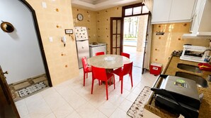 Cozinha compacta com mesa redonda, cadeiras vermelhas, geladeira e janela com cortinas.