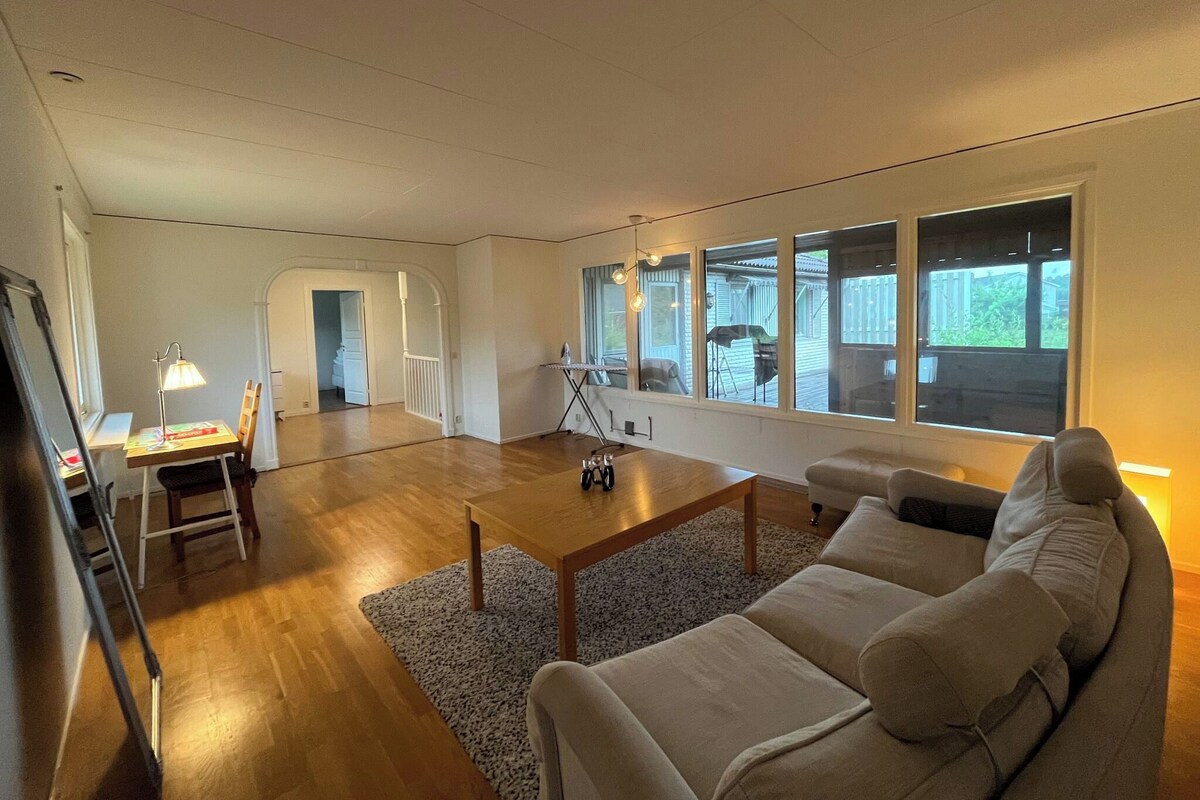 A spacious living room with a wooden coffee table, a sofa, and a desk with a lamp. There are large windows providing natural light and a view of the outside.