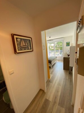 A narrow hallway leading to a bedroom with a bed and a desk, a framed picture on the wall, and a balcony with a view. - The Oyat of the Basin (Gujan-Mestras)