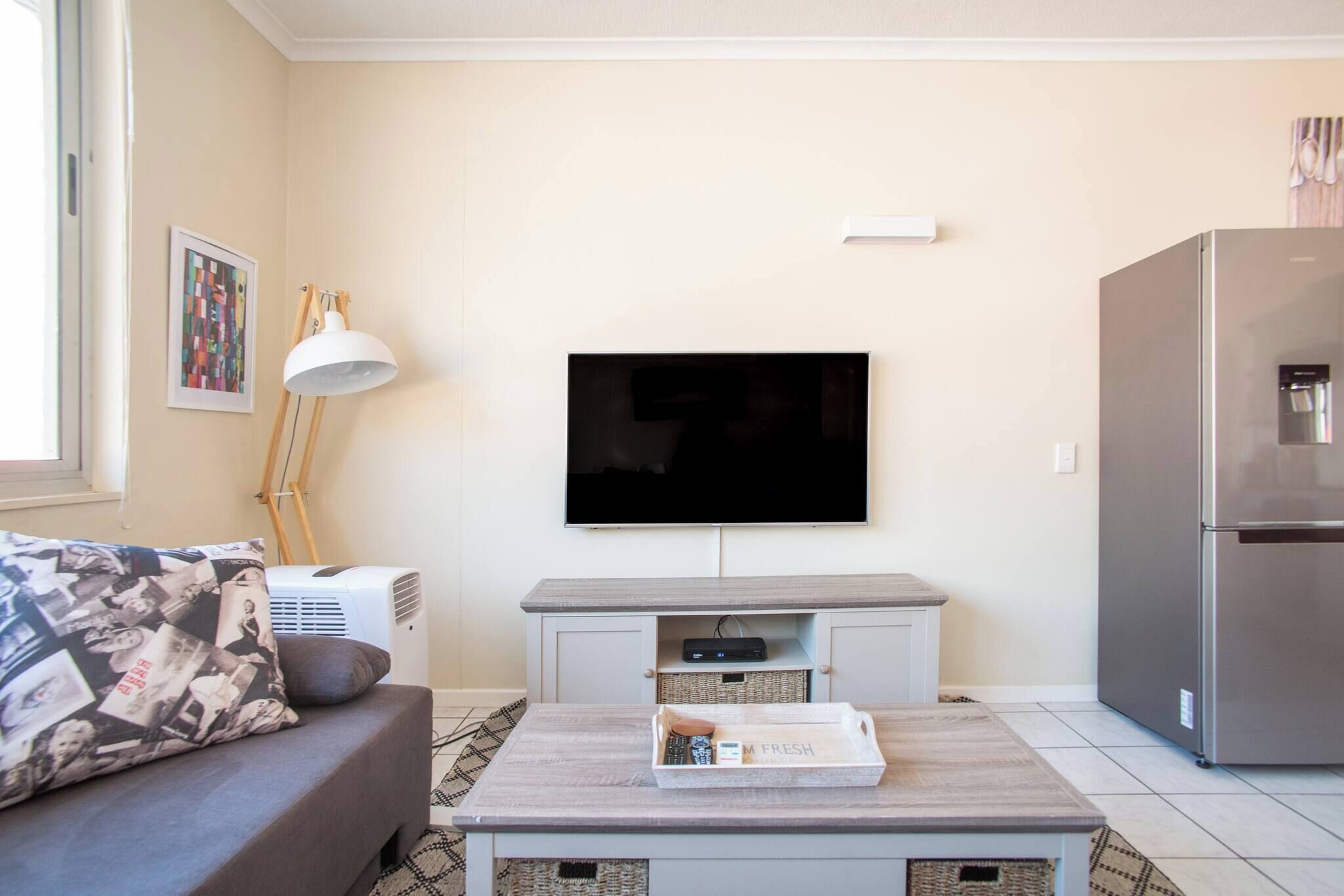 A living room with a flat-screen TV mounted on the wall, a grey sofa with a patterned throw pillow, a coffee table with a tray, and a modern refrigerator.