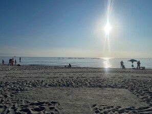 Ein Strand mit Fußabdrücken, die zum Wasser führen, ein klarer Himmel und Boote in der Ferne.