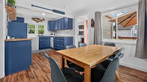 Cozinha compacta com armários azuis, uma mesa de jantar de madeira com cadeiras e vista para o exterior pela janela.