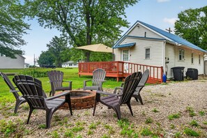 Een terras met Adirondack-stoelen, een vuurkuil en een huis met een blauw dak op de achtergrond.