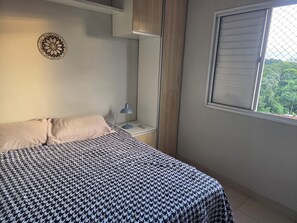 A bedroom with a bed, a nightstand, a lamp, and a window with a view of greenery. - Apartment with full air conditioning near Mangueirão (Belém)