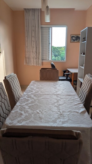 A dining area with a table and chairs, a window with blinds, a bookshelf, and a pendant light. - Apartment with full air conditioning near Mangueirão (Belém)
