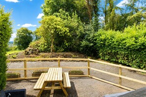 A wooden picnic table with benches, surrounded by a wire fence, set in a garden with dense greenery and trees. - Thwaite Hill Barn, Thornthwaite -  with garden, parking and wood burning stove (Thornthwaite)