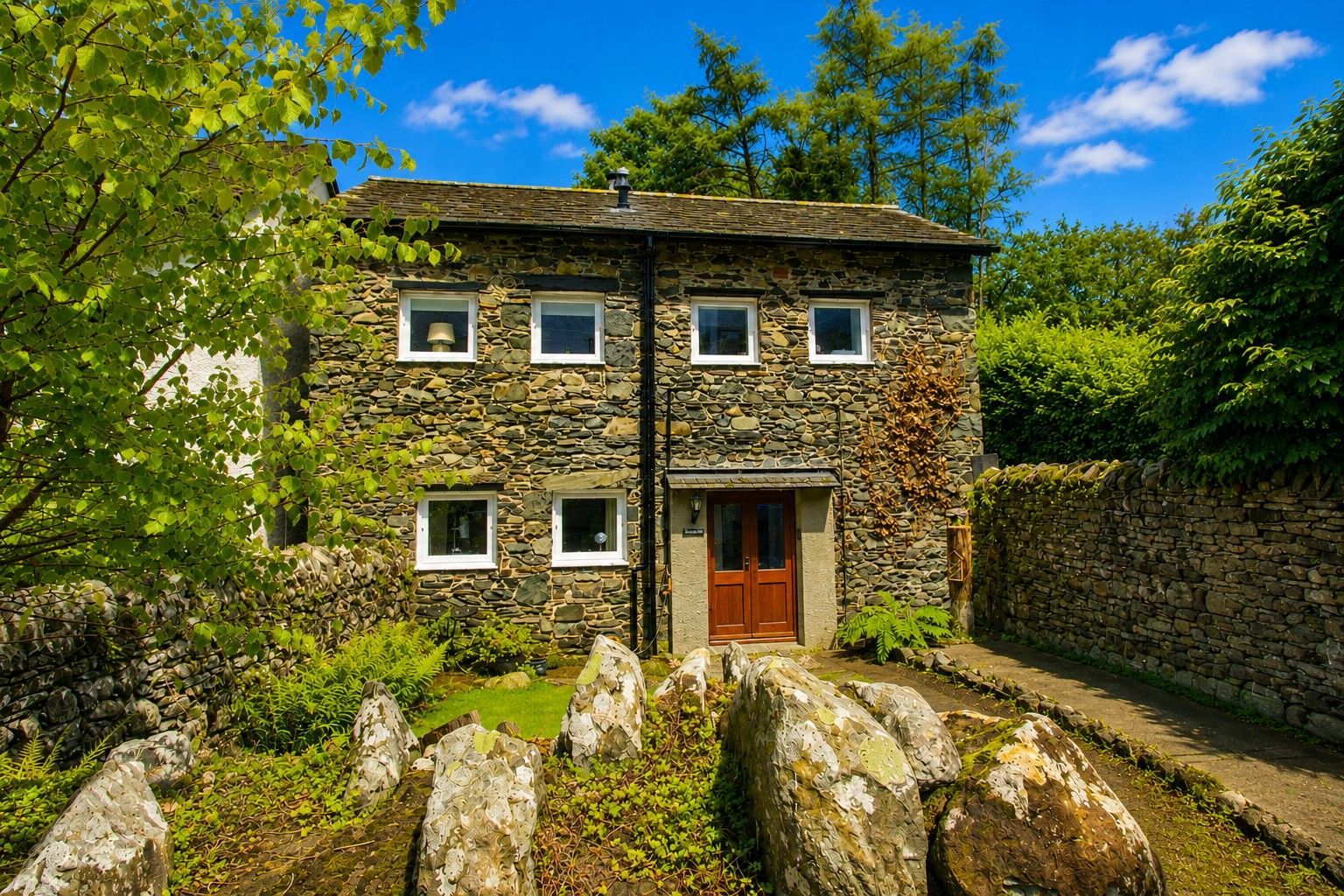A stone house with a central door and windows, surrounded by greenery and a stone wall.