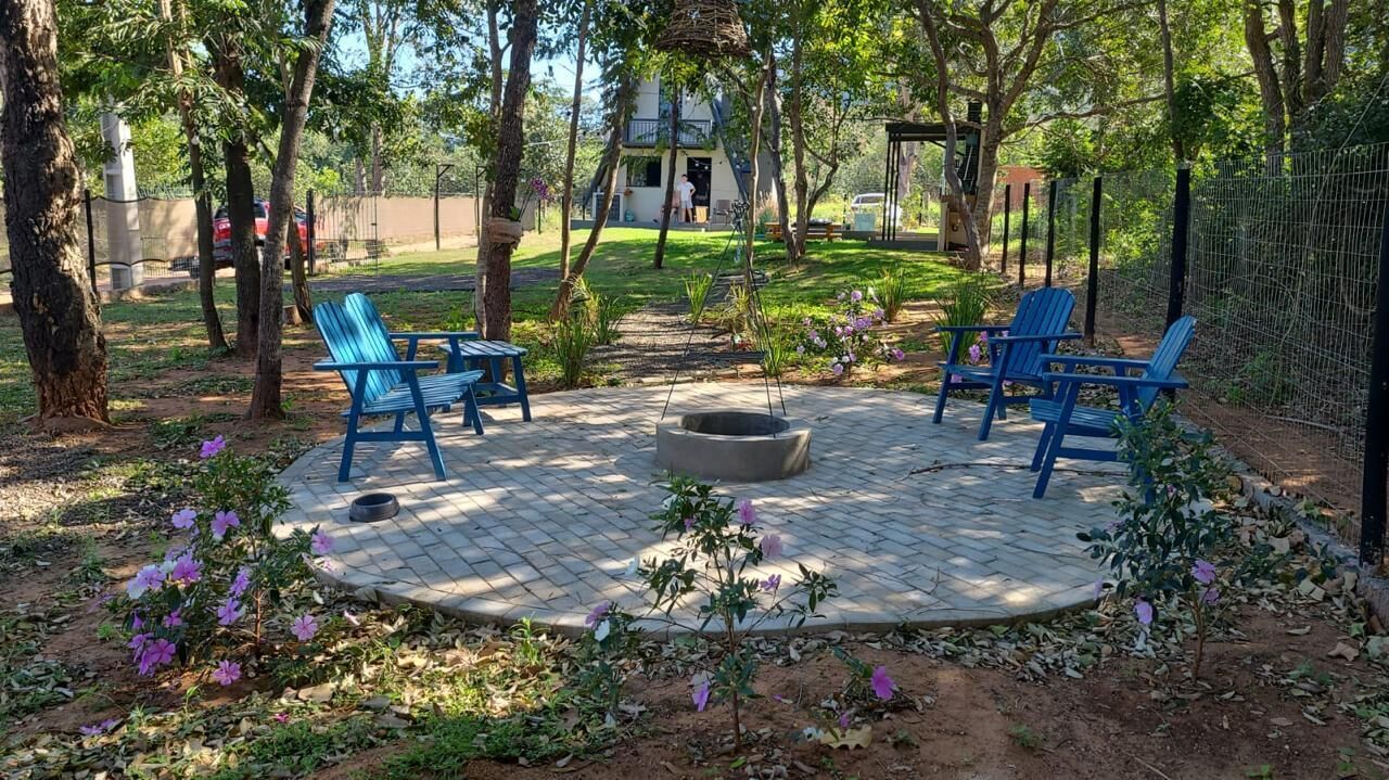 Un'area salotto all'aperto con un braciere circolare, circondata da sedie blu, in un giardino curato con alberi e piante fiorite.