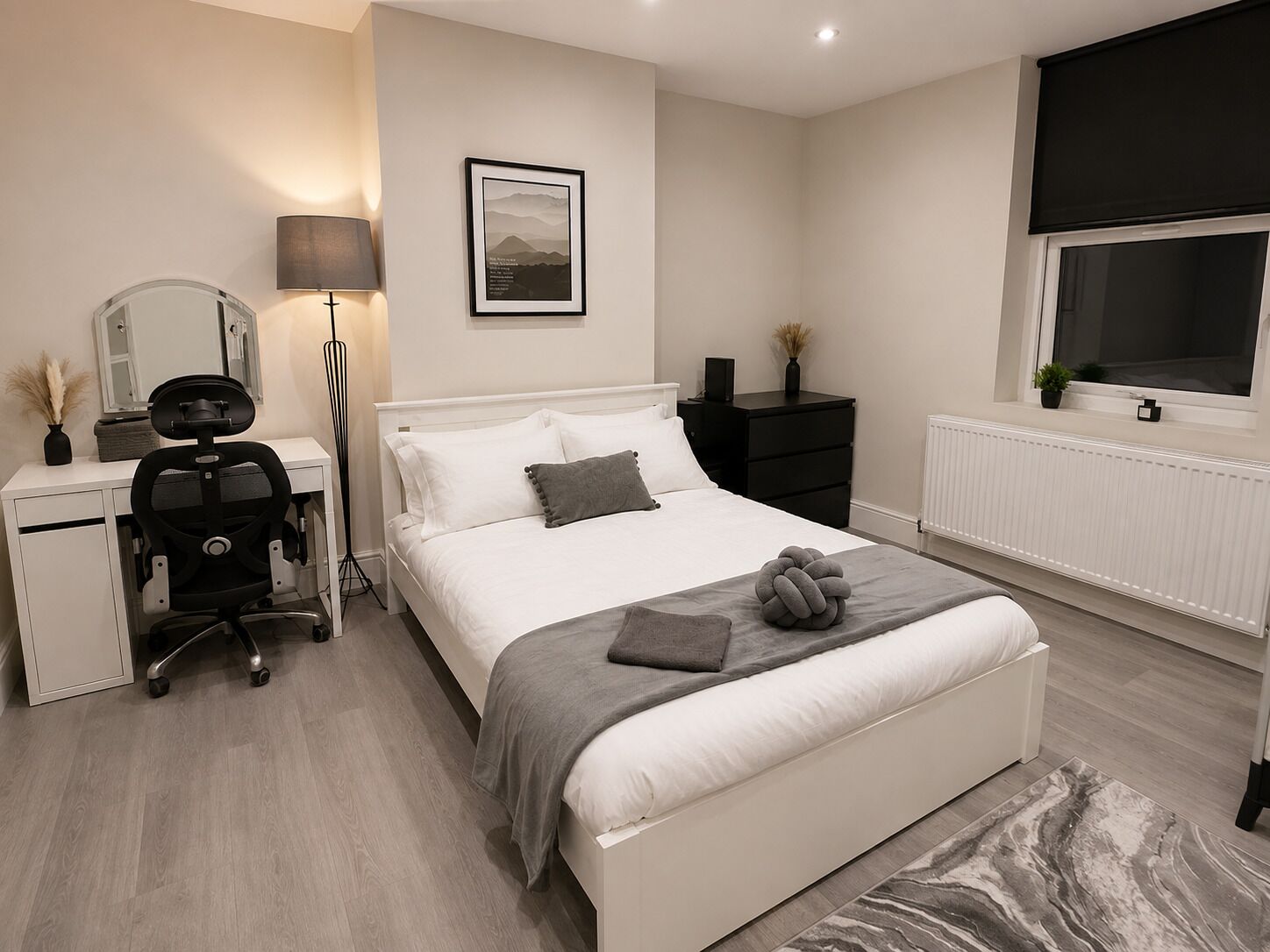 Een slaapkamer met een bed, nachtkastje, lamp, bureau, stoel en een lijst aan de muur.