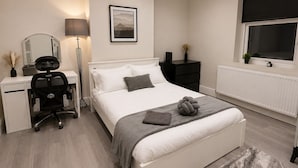 Un dormitorio con cama, mesita de noche, lámpara, escritorio, silla y un cuadro en la pared.