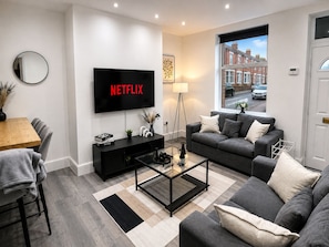 A modern living room with a flat-screen TV, a sofa, a coffee table, and a dining area. - Cosy Modern 2BR Leeds City Stay | Free Parking 1000mbps Wi-Fi (Leeds)