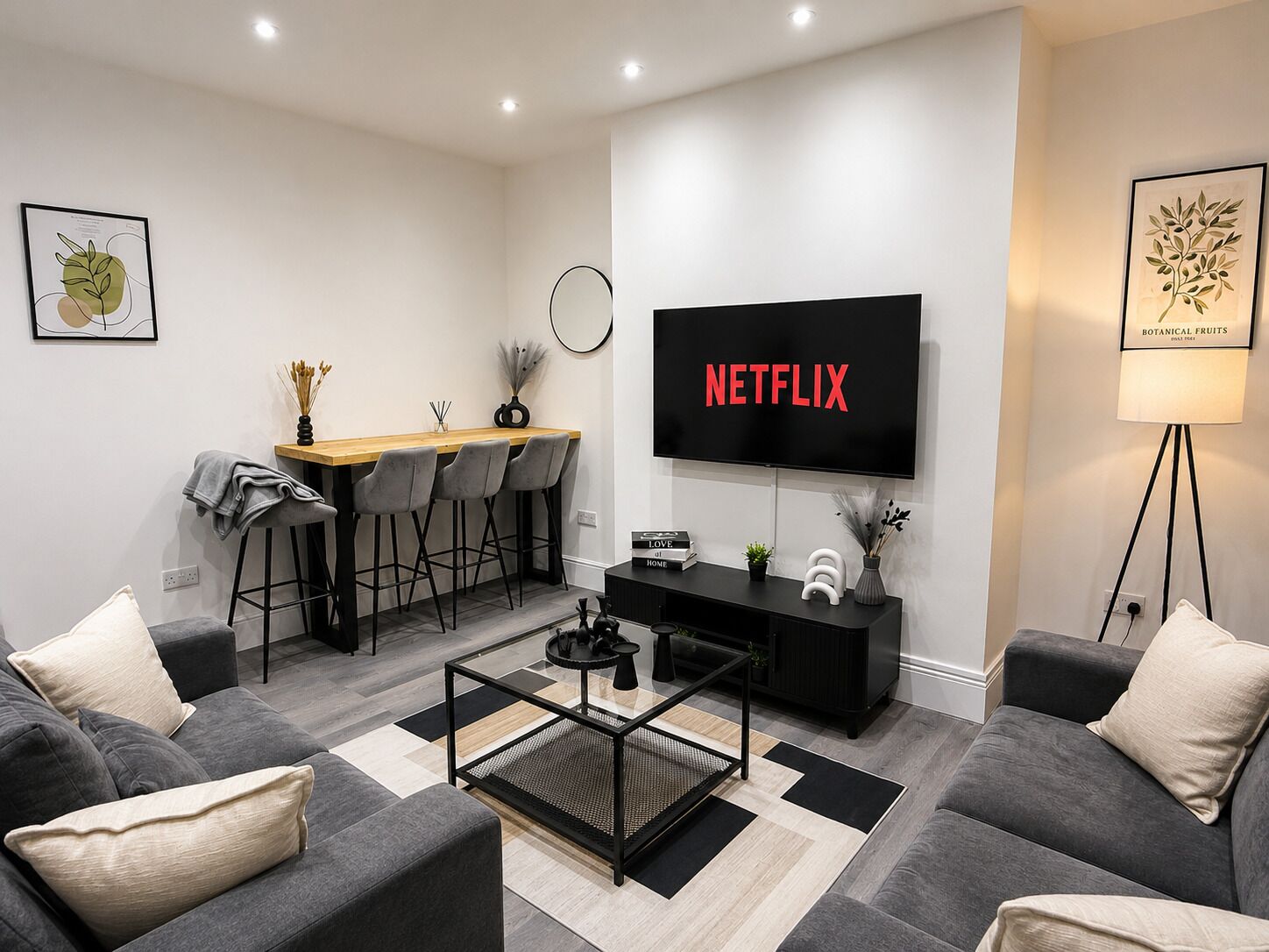 Een moderne woonkamer met een flatscreen televisie, grijze banken, een salontafel en een eethoek met barstoelen.
