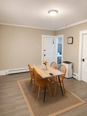 Uma sala de jantar com uma mesa de madeira, quatro cadeiras, um vaso com flores, um relógio na parede e um aquecedor branco.