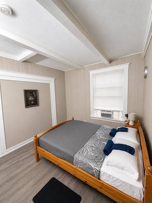 A bedroom with a wooden bed, a window with blinds, a wall-mounted air conditioner, and a framed picture.
