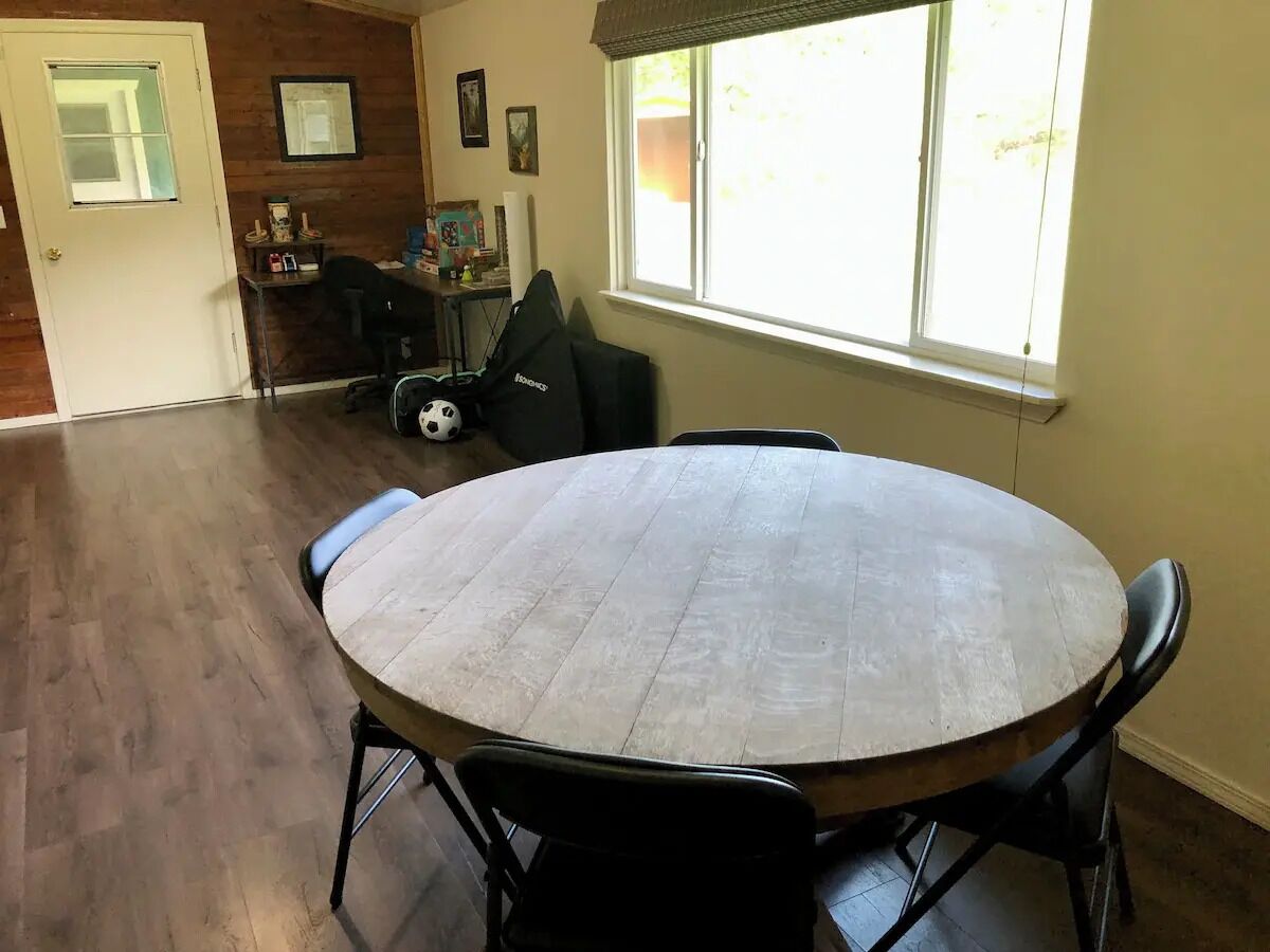 Een kamer met een ronde houten tafel, zwarte stoelen, een raam met jaloezieën en een deur naar een andere kamer.