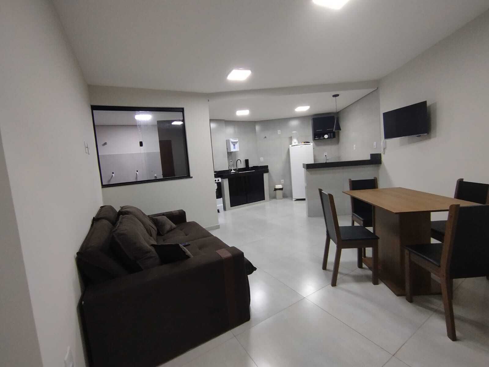 A modern living room with a sofa, a dining table with chairs, a kitchen area with a refrigerator, and a TV mounted on the wall.