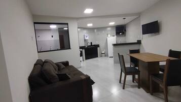 Una sala moderna con un sofá, una mesa de comedor con sillas, una zona de cocina con refrigerador y un televisor montado en la pared.