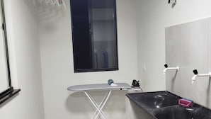 Una cocina compacta con ventana, un tendedero, una tabla de planchar, un lavamanos y un cubo de basura.