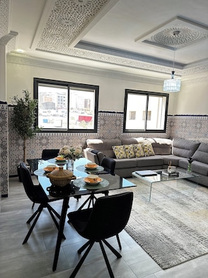 A modern living room with a glass coffee table, a sofa, and a dining area with chairs and a table set for two. - Elegant Moroccan Apartment | 2 Bedrooms & 2 Bathrooms (Tanger)