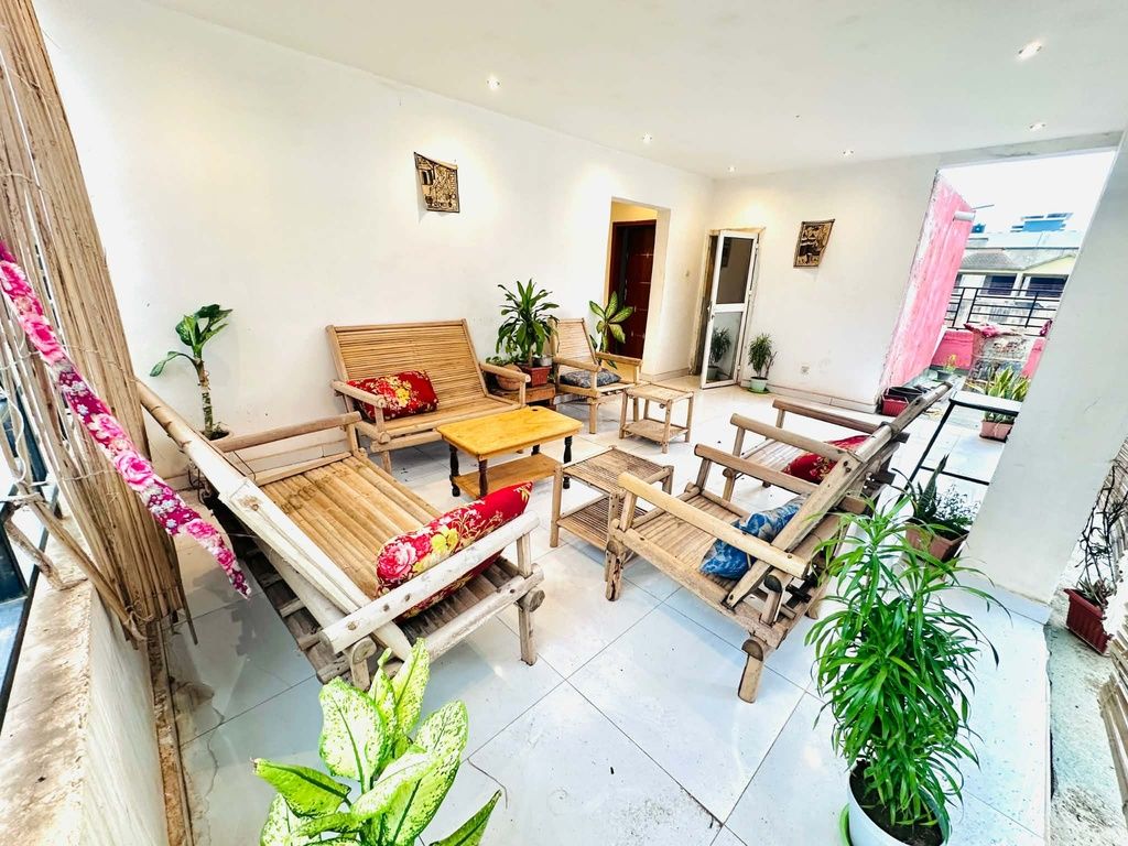 Een ruime woonkamer met een houten zithoek, een salontafel en meerdere stoelen. Er staan verschillende potplanten en er is uitzicht op buiten door een open deur.