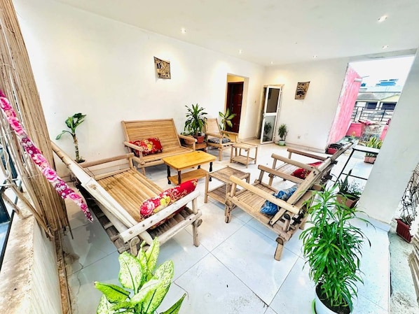 A spacious living area with a wooden sofa set, a coffee table, and multiple chairs. There are several potted plants and a view of the outside through an open door.