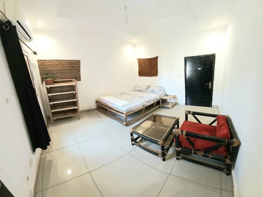 Un espacio habitable compacto con una cama, una mesita, una silla, una estantería con una planta y una puerta.