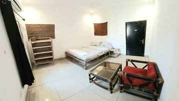 Un espacio habitable compacto con una cama, una mesita, una silla, una estantería con una planta y una puerta.