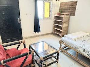 A room with a bed, a table, a chair, a window with a curtain, and a shelf with a plant.