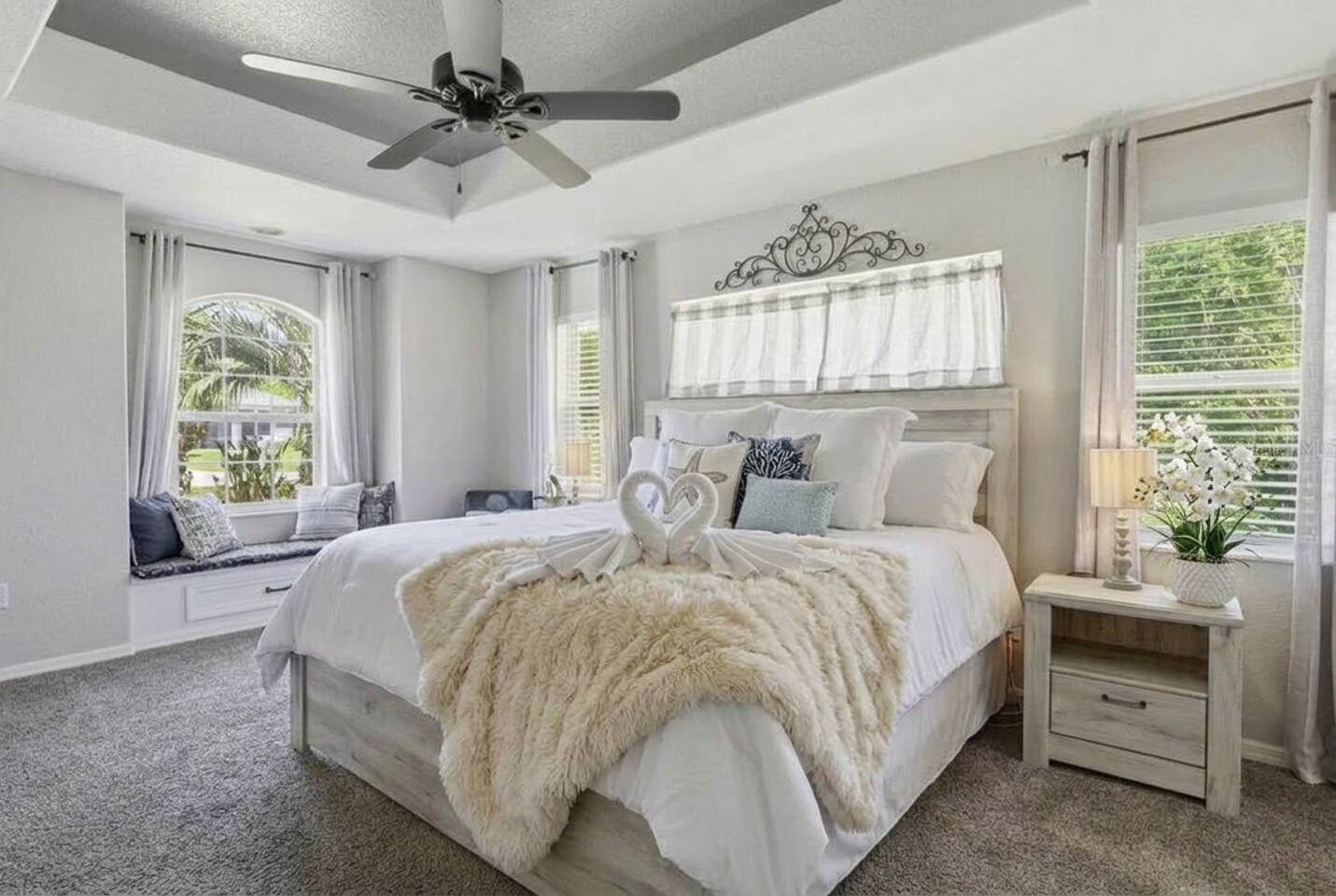 A bedroom with a large bed, a ceiling fan, a window seat, and a bedside table with a lamp.