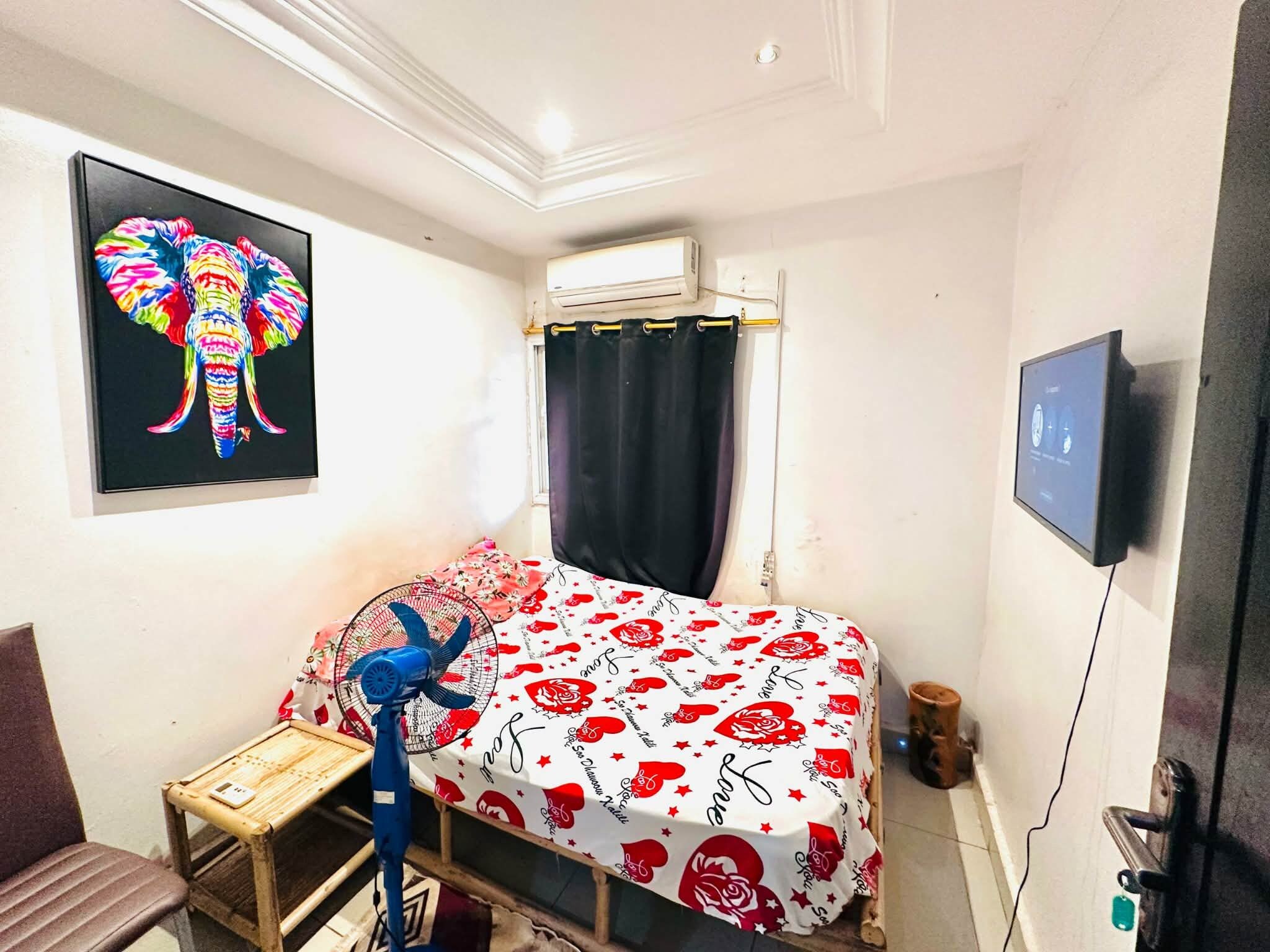 A bedroom with a bed, a small table, a chair, a fan, a TV, and a window with curtains.