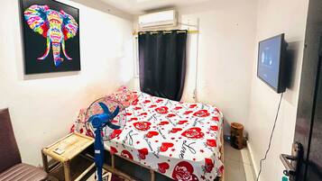 A bedroom with a bed, a small table, a chair, a fan, a TV, and a window with curtains.