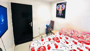 Ett sovrum med en säng, en dörr, en väggmonterad TV, en fläkt och en färgstark målning av en elefant.