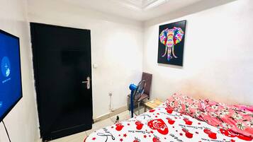 A bedroom with a bed, a door, a wall-mounted TV, a fan, and a colorful elephant painting.