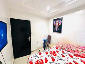 A bedroom with a bed, a door, a wall-mounted TV, a fan, and a colorful elephant painting. - Room-beautiful Room With Private Toilet (Bamako)