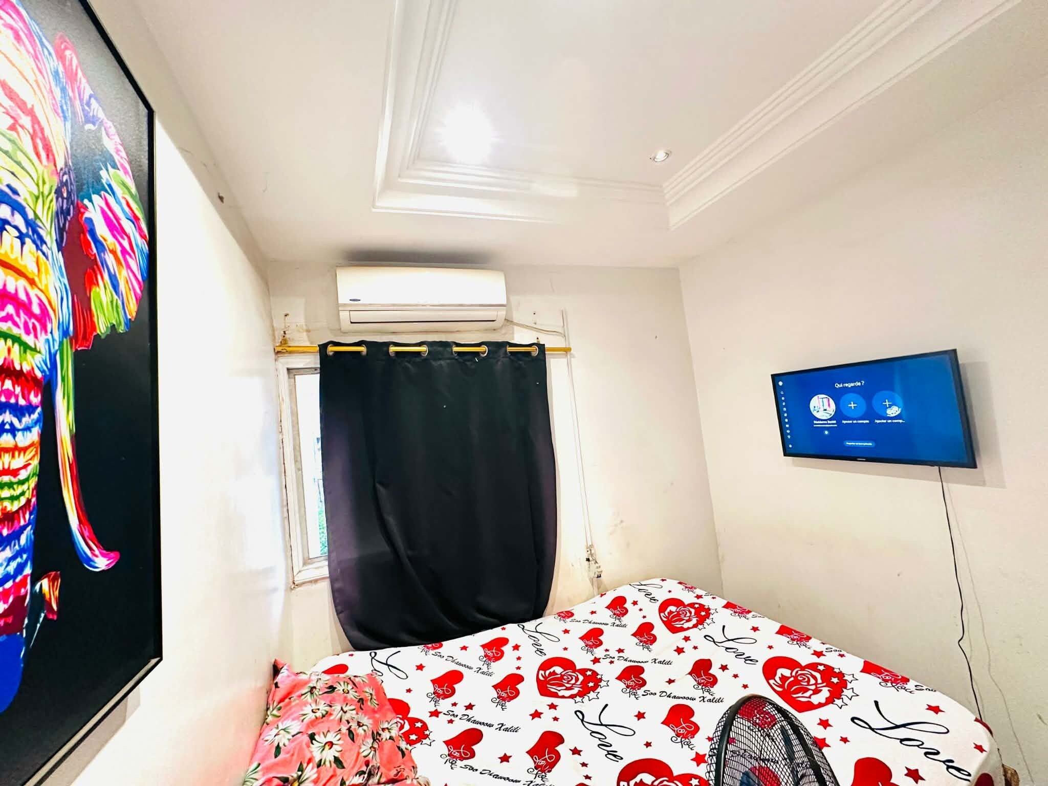 A bedroom with a bed, a TV, an air conditioning unit, and a window with a curtain.