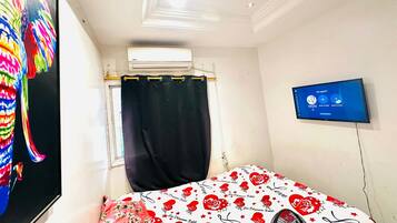 A bedroom with a bed, a TV, an air conditioning unit, and a window with a curtain.
