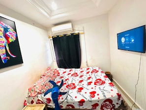 A small room with a bed, a ceiling fan, a TV, and a wall-mounted air conditioner. - Room-beautiful Room With Private Toilet (Bamako)
