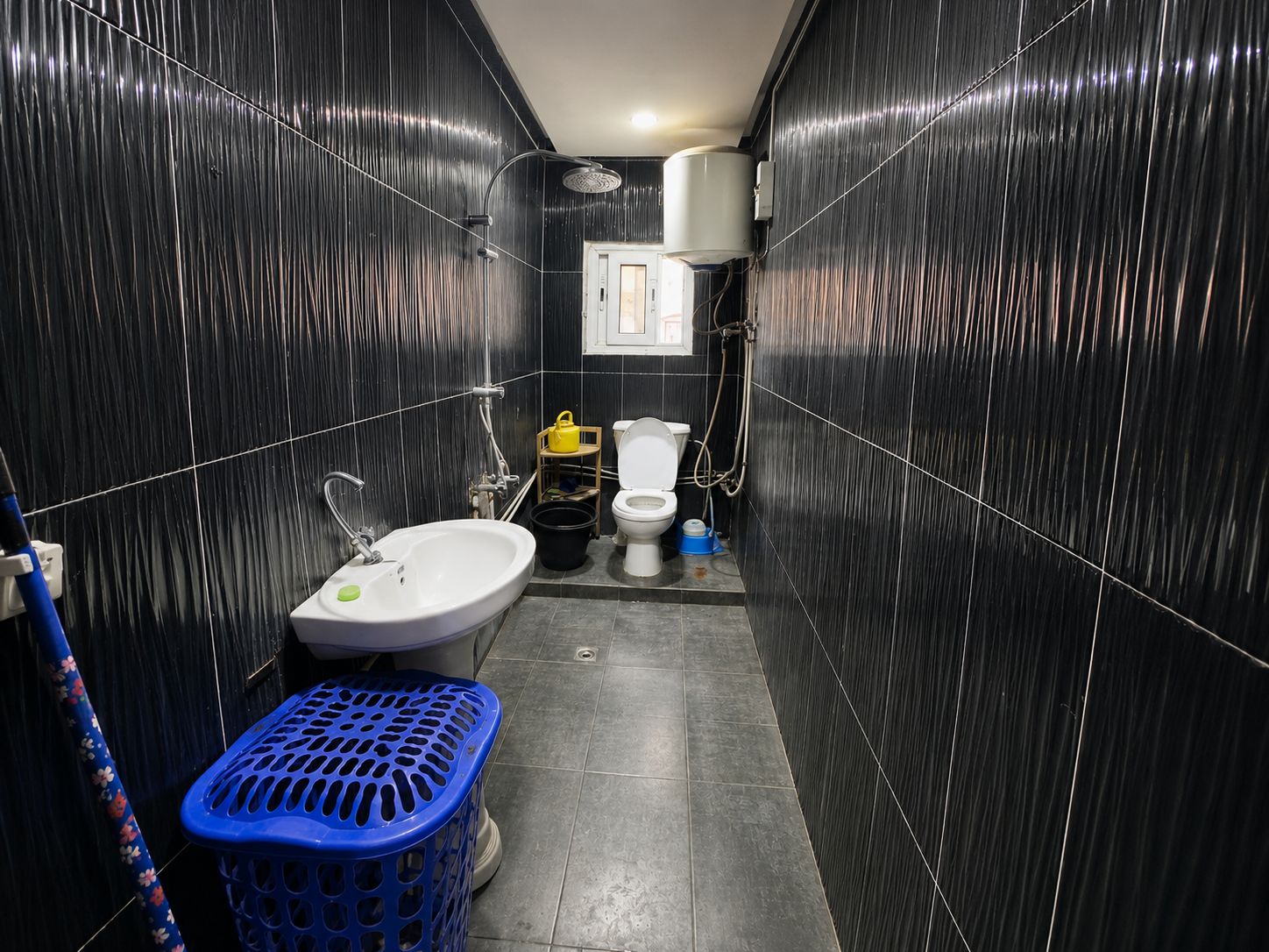 Une petite salle de bain avec un lavabo blanc, des toilettes et une douche. On y trouve un panier à linge bleu et un sac jaune sur le sol.