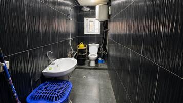 Une petite salle de bain avec un lavabo blanc, des toilettes et une douche. On y trouve un panier à linge bleu et un sac jaune sur le sol.