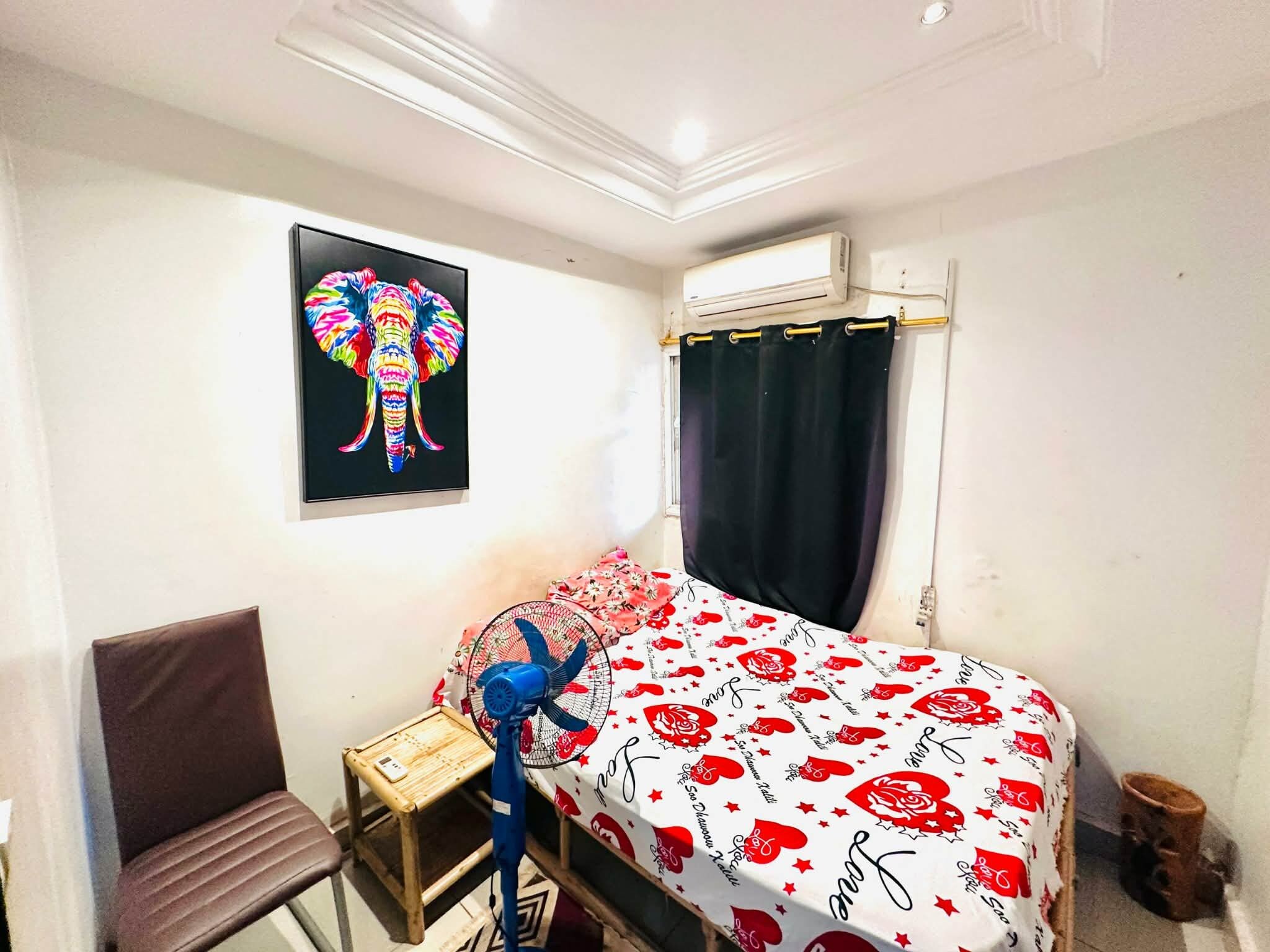 A small, well-lit room with a bed, a chair, a small table, and an air conditioning unit.