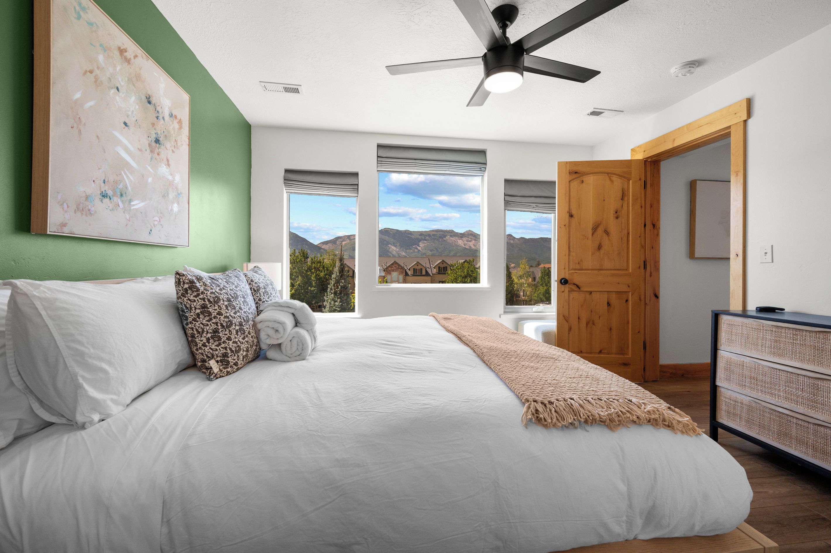 A bedroom with a bed, pillows, a blanket, a ceiling fan, and a view of mountains through windows.