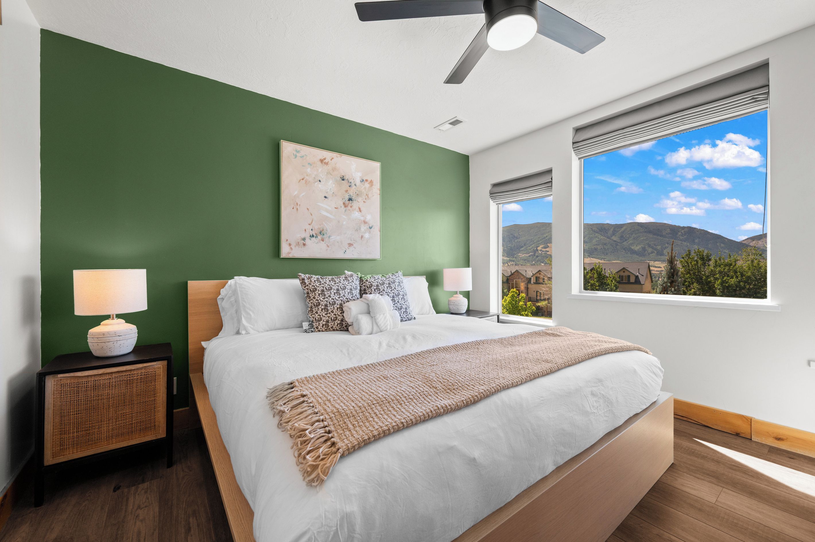 A bedroom with a bed, nightstands, a ceiling fan, a window with a mountain view, and a wall painting.