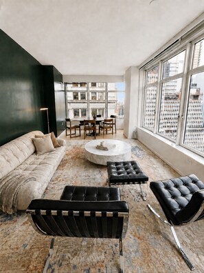 A modern living room with a sofa, two black ottomans, a coffee table, and a dining area with chairs and a table. - FiDi High-Rise · Skyline Views · Luxury Amenities (New York)